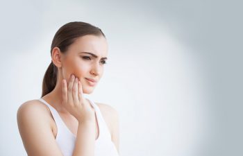 A woman in a white tank top touches her cheek and appears to be in discomfort, possibly experiencing tooth or jaw pain, against a plain light background.